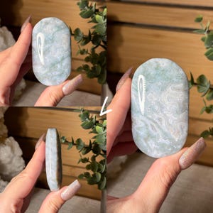 May include: A polished, oval-shaped stone with a marbled pattern of white and pale green. The stone is held in a hand, showcasing its smooth surface and natural variations. The stone's edges are rounded, and the overall aesthetic is organic and calming.