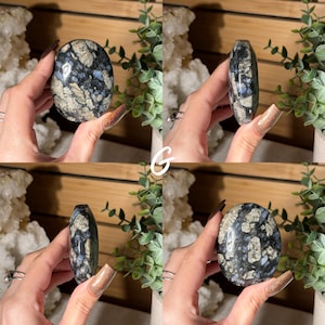 May include: A polished, oval-shaped stone with a dark blue and gray mottled pattern, accented with lighter beige and blue inclusions. The stone is held in a hand, showcasing its smooth, rounded form. The stone's surface reflects light, highlighting its unique mineral composition.
