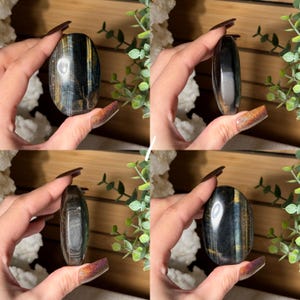 May include: A polished, oval-shaped gemstone with a mix of dark blue, gold, and black colors. The stone is held in a hand, showcasing its smooth, reflective surface. The gemstone's size is approximately 3 inches in length.