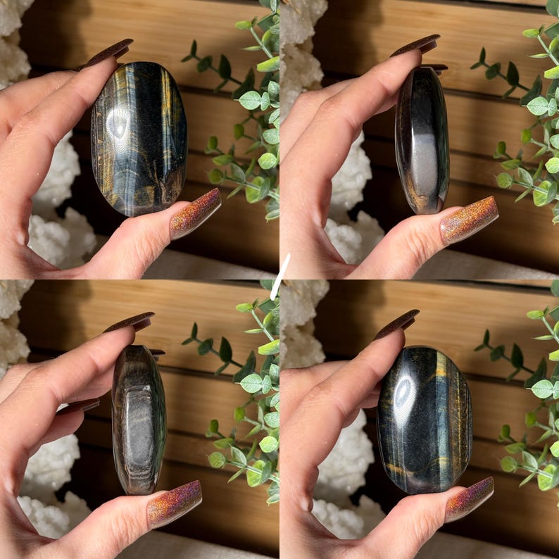 May include: A polished, oval-shaped gemstone with a mix of dark blue, gold, and black colors. The stone is held in a hand, showcasing its smooth, reflective surface. The gemstone's size is approximately 3 inches in length.