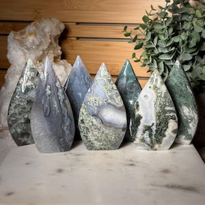Puede incluir: Una colección de siete piedras de ágata pulidas en forma de llama con patrones intrincados en tonos verde musgo, gris y blanco. Cada piedra muestra inclusiones naturales únicas, depósitos minerales en espiral y un acabado liso y brillante.