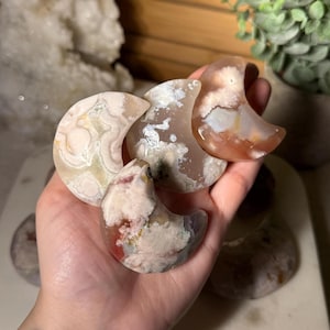 Puede incluir: Cuatro piedras pulidas en forma de media luna hechas de ágata flor. Las piedras presentan secciones translúcidas y opacas en tonos rosa suave, crema, blanco y melocotón, con intrincadas inclusiones florales visibles en todas las superficies.