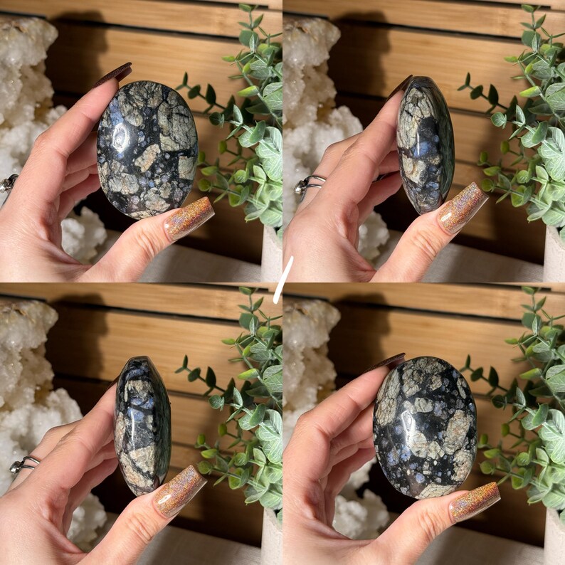May include: An oval-shaped stone with a mosaic pattern of black, gray, and blue. The polished surface of the stone reflects light, highlighting the intricate details of the natural design. The stone is held in a hand, showcasing its size and shape.
