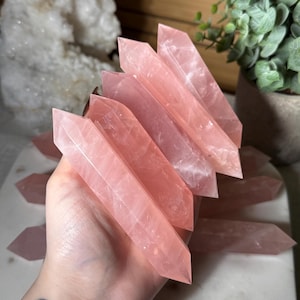 May include: A collection of pink rose quartz crystal wands. The crystals are a soft pink color and have a polished, smooth surface. The wands are pointed at both ends and are held in a hand. The background includes a white crystal and greenery.