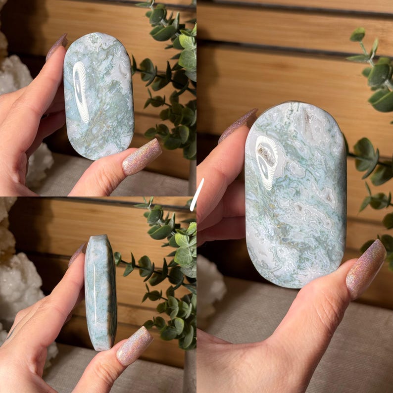 May include: A polished, oval-shaped stone with a marbled pattern of pale blue, white, and gray. The stone is held in a hand, showcasing its smooth surface and natural color variations. The stone's shape is rounded at the top and bottom.