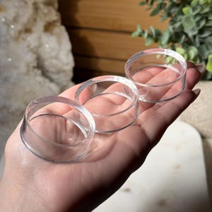 May include: Three clear, circular crystal display stands. Each stand is made of transparent acrylic and has a raised, circular rim. The stands are designed to hold small crystals or other decorative items. The stands are held in a person's hand.