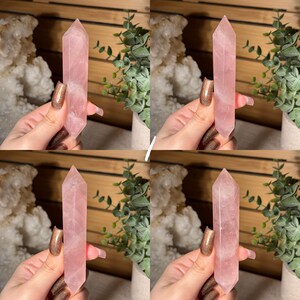 May include: Four rose quartz crystal points, each held by a hand. The crystals are a soft pink color and have a pointed, elongated shape. The background includes greenery and a white, textured object.
