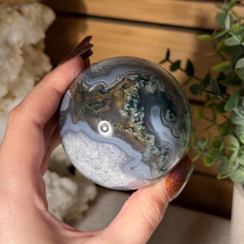 May include: A polished moss agate sphere. The sphere features a mix of colors, including white, gray, brown, and green, with a swirling pattern. The sphere is held in a hand, with a wooden background and greenery in the background.