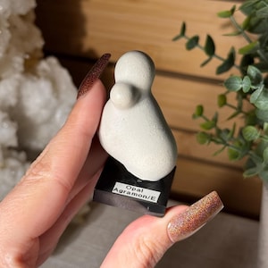 May include: A smooth, off-white, rounded stone sculpture with a smaller protrusion on its side, mounted on a square black base. A white label on the front of the base features the black printed text "Opal Agramon/E" in two lines.