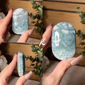 May include: A polished, oval-shaped stone with a marbled appearance. The stone features a mix of white and green hues, with intricate patterns resembling moss or veins. The stone is held up in front of a wooden background.
