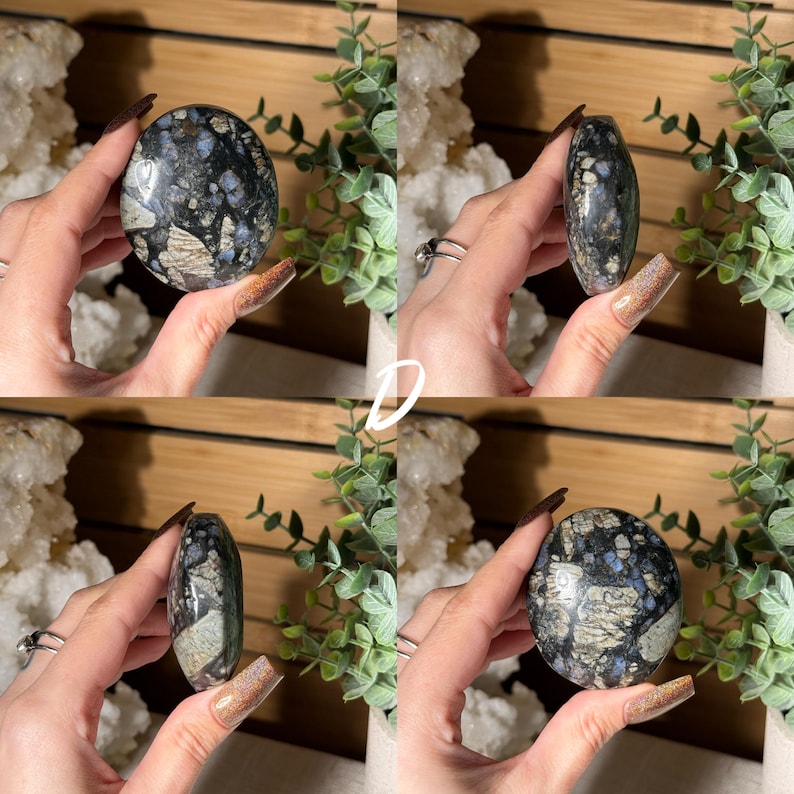 May include: Four views of a polished, oval-shaped gemstone. The stone features a mix of dark blue, gray, and white patterns. The gemstone is held in a hand, showcasing its shape and texture. The background includes greenery and a wooden surface.