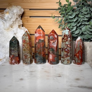Puede incluir: Seis torres de cristal pulido dispuestas en fila, con patrones variados de inclusiones minerales en rojo intenso, marrón tierra y verde musgo. Cada punta de piedra tiene una superficie lisa y facetada con formaciones de color únicas.