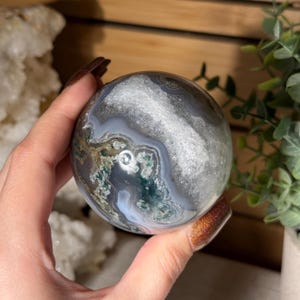 May include: A polished, spherical gemstone with a swirling pattern of gray, white, and green hues. The orb-shaped stone is held in a hand, showcasing its natural beauty and intricate details. This decorative item is a unique addition to any collection.
