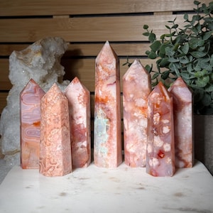 May include: A collection of seven pink and orange agate crystal obelisks. The crystals have a variety of patterns and colors, including white, orange, and pink. The obelisks are standing on a white surface.