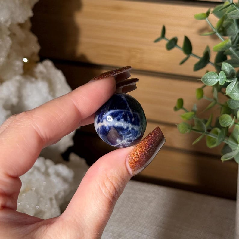 K&ouml;nnte beinhalten: Eine kleine, runde Sodalit-Kugel, gehalten zwischen Fingern. Der polierte Stein weist eine tiefblaue Farbe mit wei&szlig;en und grauen Adern auf. Die Kugel hat einen Durchmesser von etwa 3,8 cm und zeigt nat&uuml;rliche Muster und Texturen.