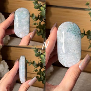 May include: A polished, oval-shaped stone with a marbled pattern of white, light blue, and green hues. The stone is held in a hand, showcasing its smooth surface and natural variations. The stone's shape is elongated, with rounded edges.