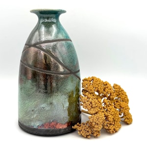 May include: A ceramic vase with a narrow neck and a wide base, featuring a glossy glaze in shades of teal, copper, and dark brown with a fine crackle pattern. The surface is decorated with dark, sweeping diagonal lines and dried yellow flowers.