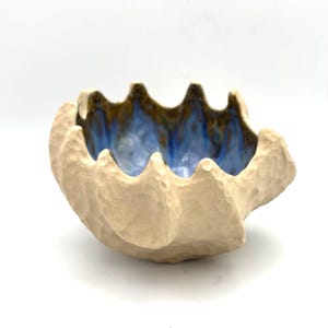 May include: A ceramic bowl with a textured, matte beige exterior featuring a scalloped, uneven rim. The interior of the bowl is glazed in a glossy, vibrant blue color that transitions into dark brown tones near the top edge of the vessel.