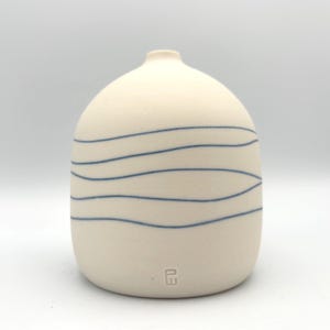 Pode incluir: Um vaso de cerâmica creme com corpo arredondado e pescoço estreito. O vaso é adornado com linhas azuis horizontais em forma de onda. Na base, há uma pequena marca estampada. Um item decorativo.