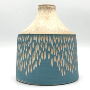 May include: A ceramic vase with a teal blue base and a cream-coloured top. The blue section features a pattern of vertical, teardrop-shaped cutouts. The vase has a rounded shape and a narrow neck, suitable for displaying flowers or as a decorative piece.