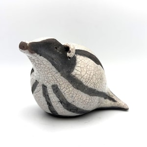 May include: A ceramic badger figurine featuring a white crackle glaze finish with bold, dark grey stripes across the body and head. The sculpture has a rounded, compact shape with a pointed snout and small ears, showcasing a distinct texture.