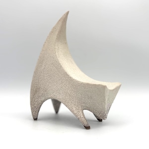 May include: A small, off-white ceramic sculpture of a four-legged creature with a tall, curved back and a pointed head. The surface features a distinct dark crackle glaze pattern throughout, with four pointed feet touching the ground.