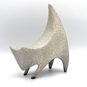 May include: A ceramic sculpture of a four-legged animal with a curved, pointed back. The surface is off-white with a dark, intricate crackle glaze pattern throughout. The four feet are dark-toned, and the overall form is abstract and angular.