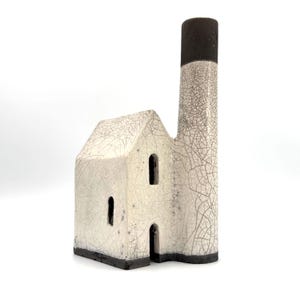 May include: A ceramic house-shaped sculpture with a tall chimney, featuring a white crackle glaze finish over the main structure and a dark brown base and chimney top. The building has three arched openings representing windows and a doorway.