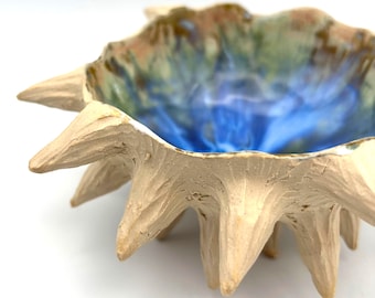 May include: A handmade ceramic bowl with a unique, spiky design. The bowl features a textured, off-white exterior and a vibrant interior with blue, green, and brown hues. The bowl's rim has a jagged, star-like shape.