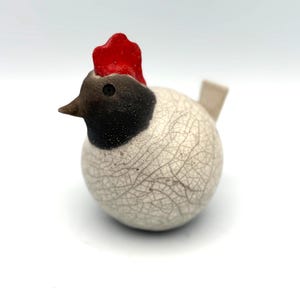 May include: A small, round ceramic chicken figurine featuring a white body with a dark crackle glaze finish, a dark brown head, a bright red comb, and a small, light-coloured tail. The bird has a pointed beak and a single visible dark eye.