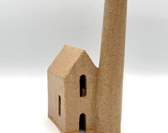 May include: A tan ceramic sculpture shaped like a small factory building with a tall, cylindrical chimney. The structure features a gabled roof, a rectangular doorway at the base, and two arched window openings on the front facade with a textured finish.