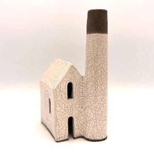 May include: A ceramic house-shaped sculpture with a tall chimney, featuring a white crackle glaze finish across the structure and a dark brown matte texture on the top of the chimney. The building has a gabled roof and two arched openings.