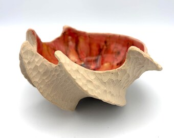 May include: A ceramic bowl with a textured, matte beige exterior featuring carved facets. The interior of the bowl is finished with a glossy, vibrant glaze in shades of red, orange, and yellow, creating a stark contrast with the raw outer surface.
