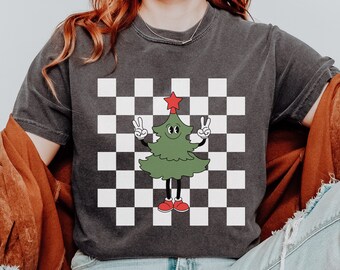 May include: A black t-shirt with a checkered pattern and a cartoon Christmas tree wearing red shoes and giving the peace sign. The tree has a red star on top.