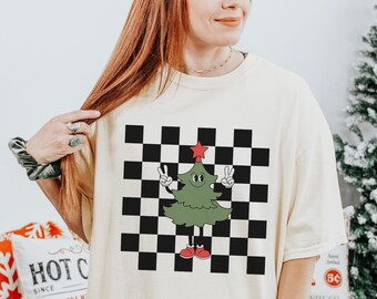 May include: A cream colored t-shirt with a black and white checkered pattern and a cartoon Christmas tree wearing red shoes. The tree is holding up two fingers in a peace sign.