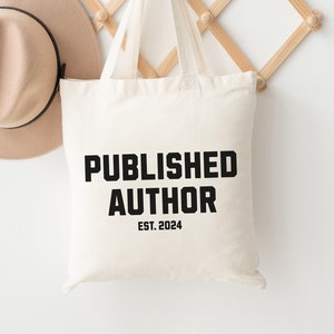 May include: A beige tote bag with the text "PUBLISHED AUTHOR EST. 2024" in black, hanging on a wooden wall rack. A beige hat with a black band is also hanging on the rack. The background is a white wall.