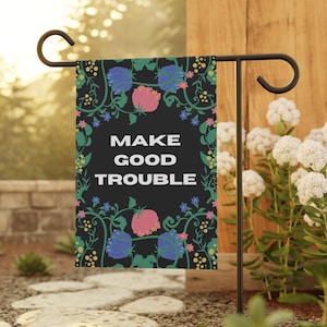 May include: A black garden flag with the words "MAKE GOOD TROUBLE" in white. The flag is decorated with pink and blue floral designs and is displayed on a black metal stand in a garden setting.