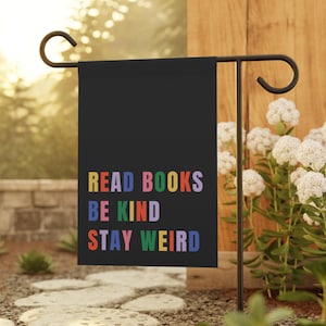 May include: A black garden flag with the words "READ BOOKS BE KIND STAY WEIRD" in colorful block letters. The flag is hanging on a black metal stand in a garden setting with white flowers and stone pavers.