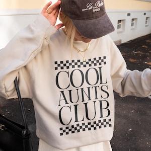 May include: A beige sweatshirt with the text "COOL AUNTS CLUB" in black, framed by a black and white checkered pattern. A brown baseball cap with "La Paris" in white is worn. A black purse and a beverage in a clear cup are also visible.