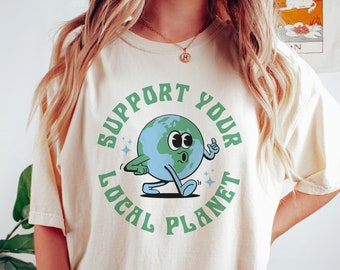 May include: A woman wearing a cream colored t-shirt with a green circle graphic that says "Support Your Local Planet". The graphic features a cartoon earth with a face and arms and legs. The woman is wearing green biker shorts.