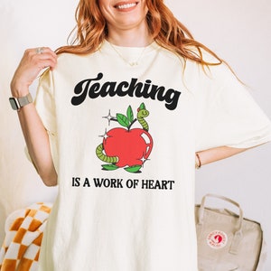 May include: A white t-shirt with the text "Teaching is a work of heart" in black. A red apple with a green worm and leaves is above the text.