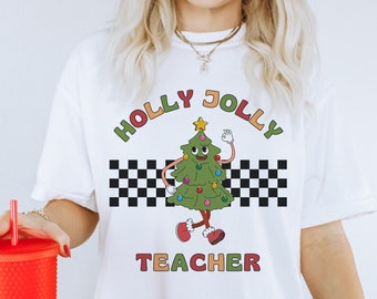 May include: White t-shirt with a cartoon Christmas tree wearing red shoes and holding a peace sign. The text "HOLLY JOLLY TEACHER" is printed on the shirt in rainbow colors.
