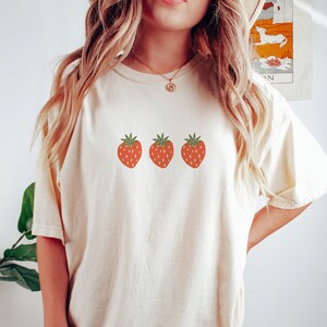 May include: A cream colored t-shirt with a graphic of three red strawberries with green leaves.