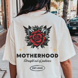 May include: White t-shirt with a red rose and wings graphic on the back. The text "MOTHERHOOD Straight out of patience EST. 2025" is printed below the graphic.