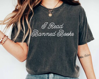 May include: A woman wearing a dark gray t-shirt with the text "I Read Banned Books" in white cursive lettering. She is wearing blue jeans and white sneakers.