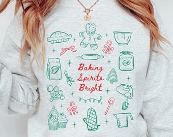 May include: A light gray sweatshirt with a festive baking-themed design. The design includes green and red illustrations of baking tools and treats, with the words "Baking Spirits Bright" in the center.