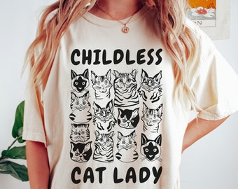 May include: A cream-colored t-shirt with the text "Childless Cat Lady" printed in black. The shirt features a graphic of multiple black and white cat faces.