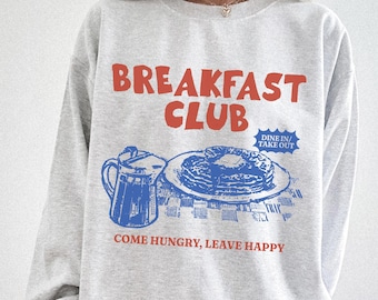 May include: A light gray sweatshirt with a retro graphic design. The design features the words "BREAKFAST CLUB" in red, along with a blue illustration of pancakes, syrup, and a pitcher. The text "COME HUNGRY, LEAVE HAPPY" is also included.