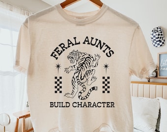 Op de afbeelding: Crèmekleurig T-shirt met de zwarte tekst "FERAL AUNTS BUILD CHARACTER". Een zwart-witte tijgerafbeelding staat in het midden van het shirt, met dambordpatronen aan weerszijden. Het shirt hangt aan een houten hanger.