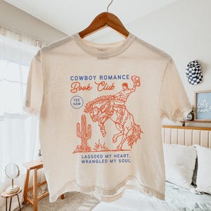 May include: Cream-colored t-shirt with a red and blue graphic. The graphic features a cowboy riding a bucking bronco, a cactus, and the text "Cowboy Romance Book Club." Additional text reads "Yee Haw" and "Lassoed My Heart, Wrangled My Soul."